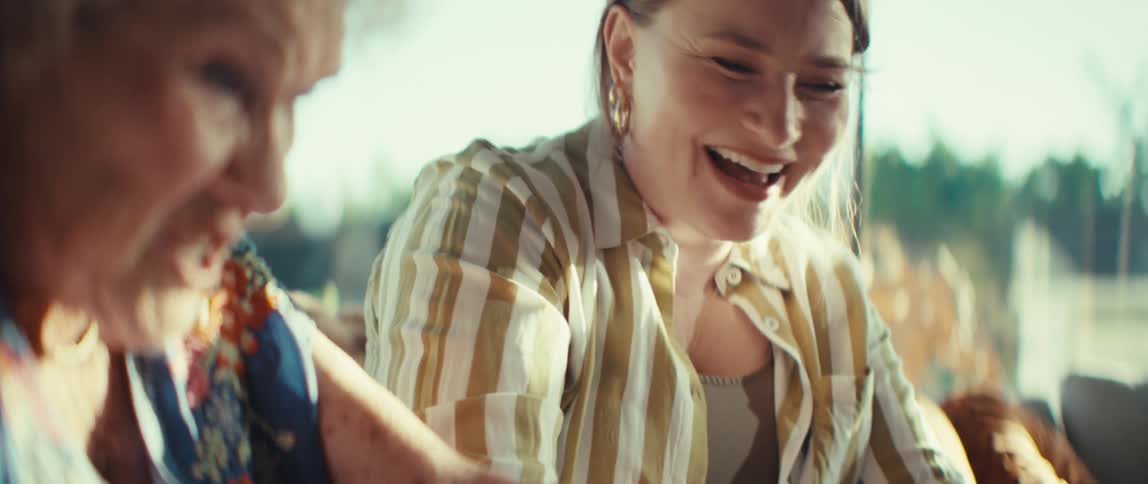 A younger woman wearing a striped shirt laughs and expresses surprise while sitting next to an elderly womanthe scene captures a closeup candid moment between the two as they engage in a lively conversation