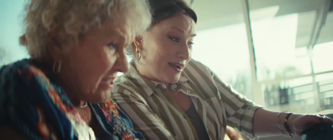 An elderly woman and a younger woman sit together indoors and use black controllers to play a video game with excited expressions while sunlight shines into the room from a nearby window