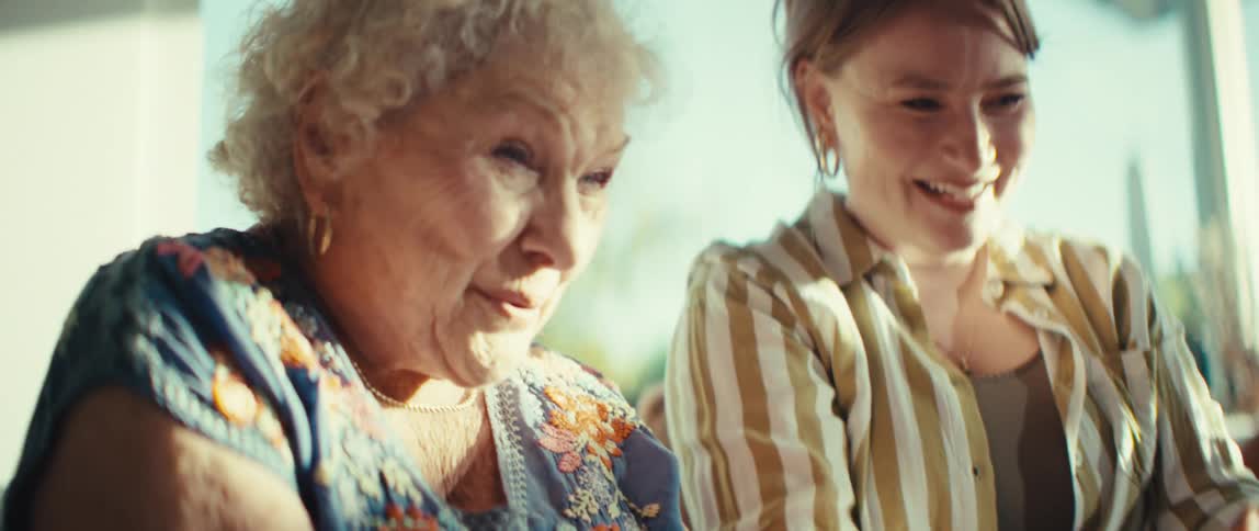 An elderly woman and a younger woman laugh and cheer while playing video games together