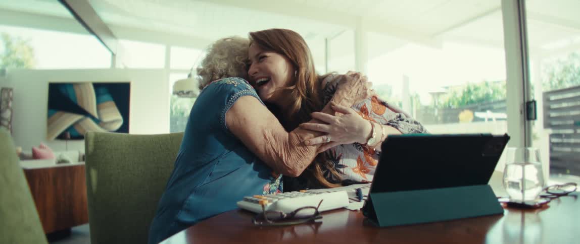 A view from a side angle shows an older woman and a younger woman sitting at a table together laughing and hugging in front of a tablet and financial documents