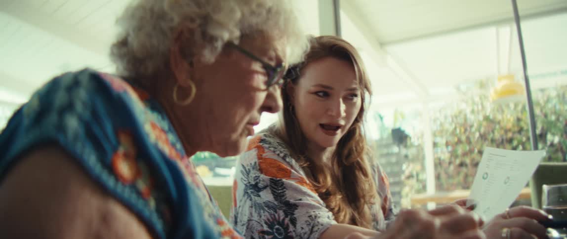 A younger woman wearing a floral shirt embraces an elderly woman while they sit together indoors with a tablet on the table and look at each other with sad expressions