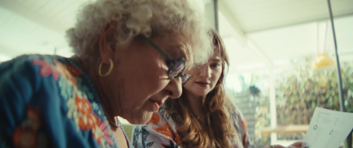 An elderly woman wearing glasses and a younger woman sit side by side at a table while looking at a document and using a calculator in a bright room