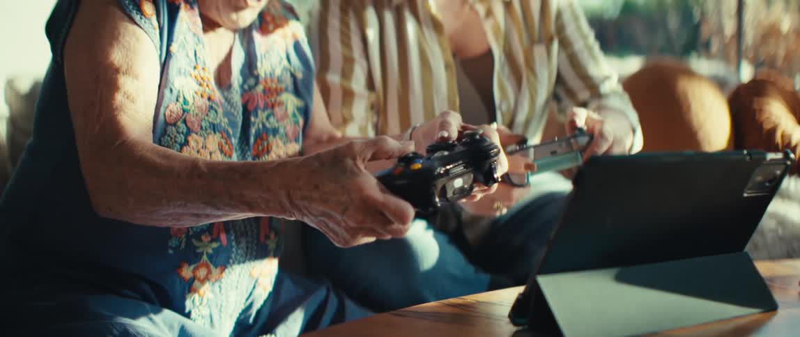 A younger woman and an elderly woman sit on a sofa and play a game using hand held controllers while looking at a tablet on a table until they both react with wide mouths and surprised expressions