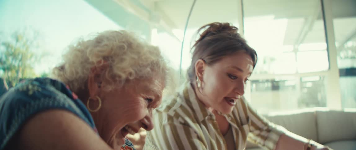 Two women one young and one elderly sitting on a couch playing a video game with controllers having fun in the living room at midday