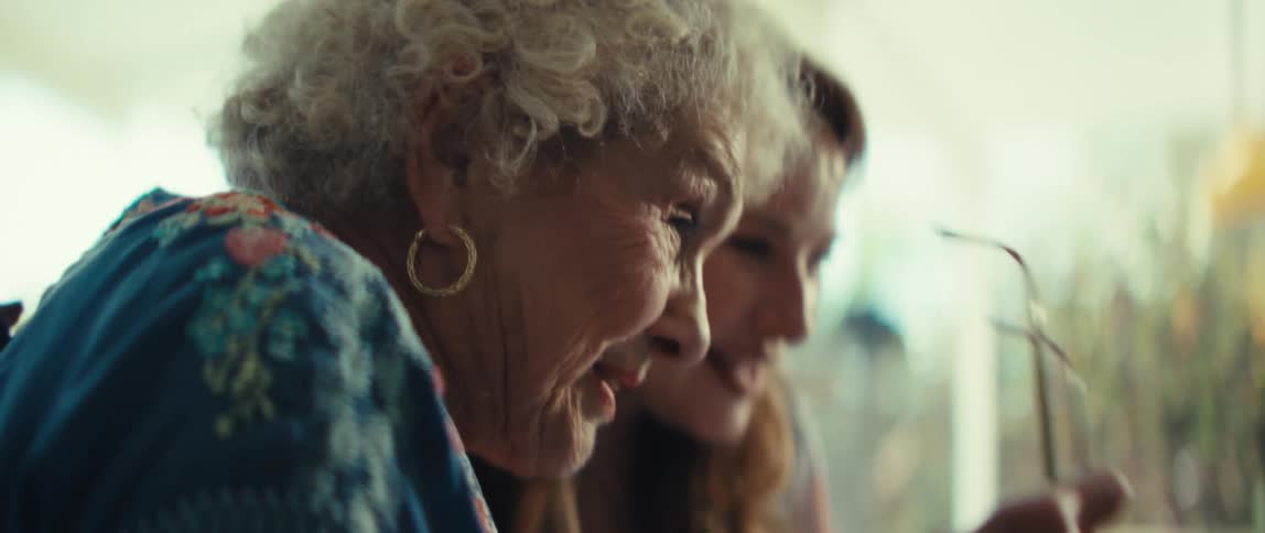 A close view of an elderly woman with white curly hair wearing gold earrings and a blue floral shirt as she talks and smiles while sitting next to a younger woman in a brightly lit indoor setting