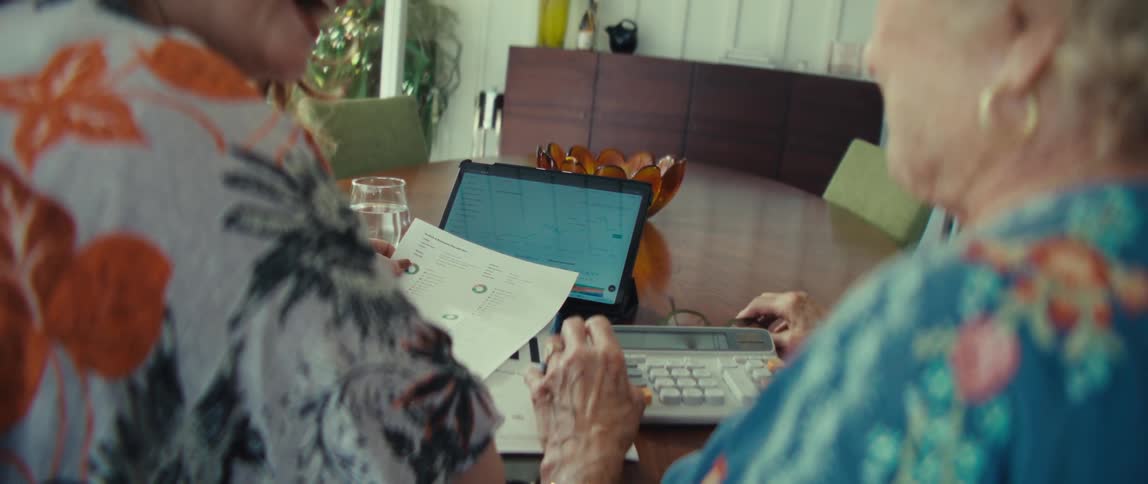 A view from behind shows two women sitting at a wooden table with a tablet and papers while an elderly woman uses a calculator and reacts with a surprised gesture