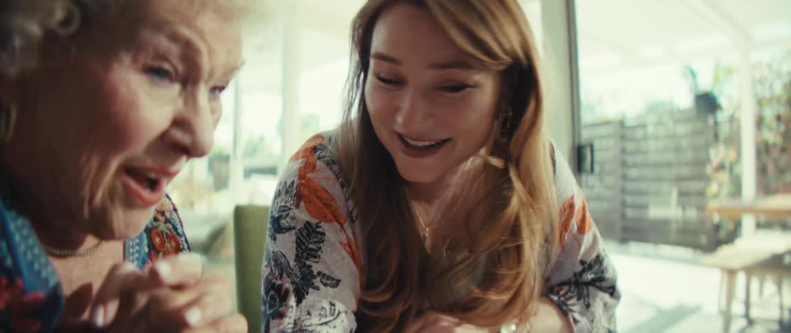 A younger woman with long brown hair smiles and points at a tablet screen while sitting at a wooden table with an elderly woman who has white curly hair and looks on with a surprised expression