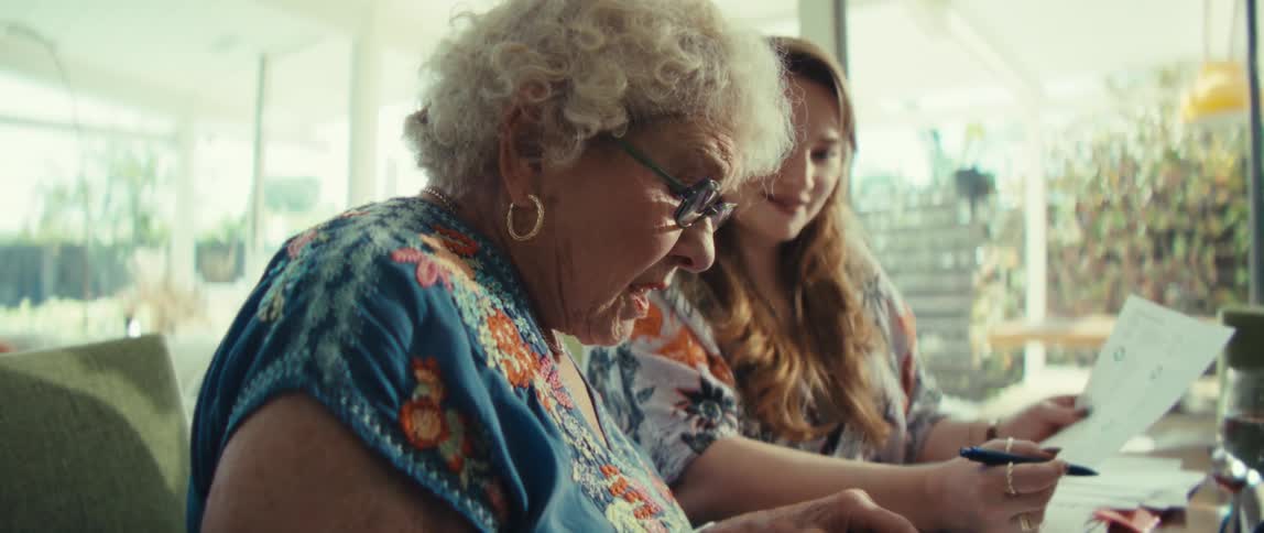A younger woman assists an elderly woman with paperwork and a digital tablet at a table