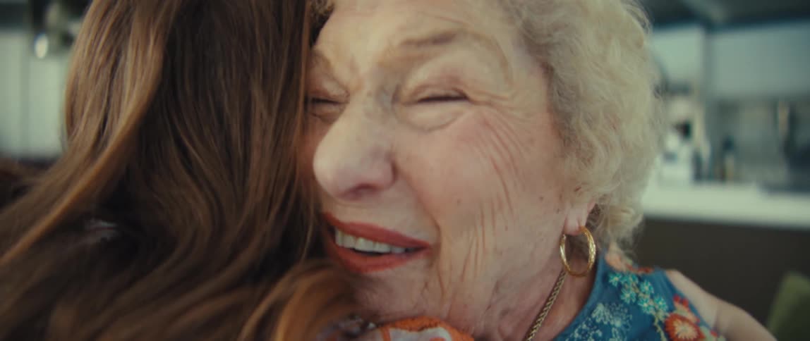 An older woman with curly white hair wearing a blue floral shirt reacts with surprise and hugs a younger woman with long brown hair in a bright kitchen during the day