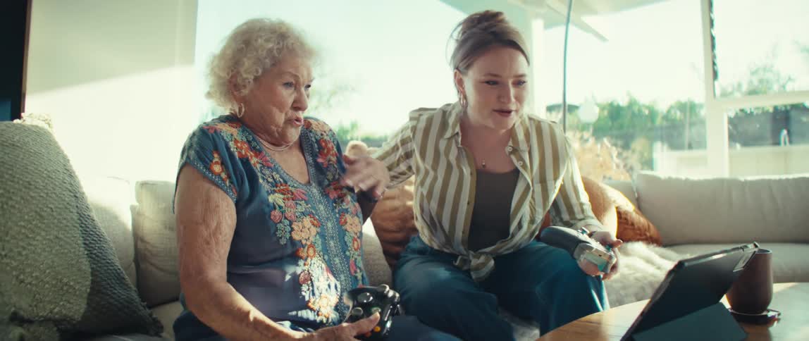 An elderly woman and a younger woman laugh and cheer while playing video games together on a couch