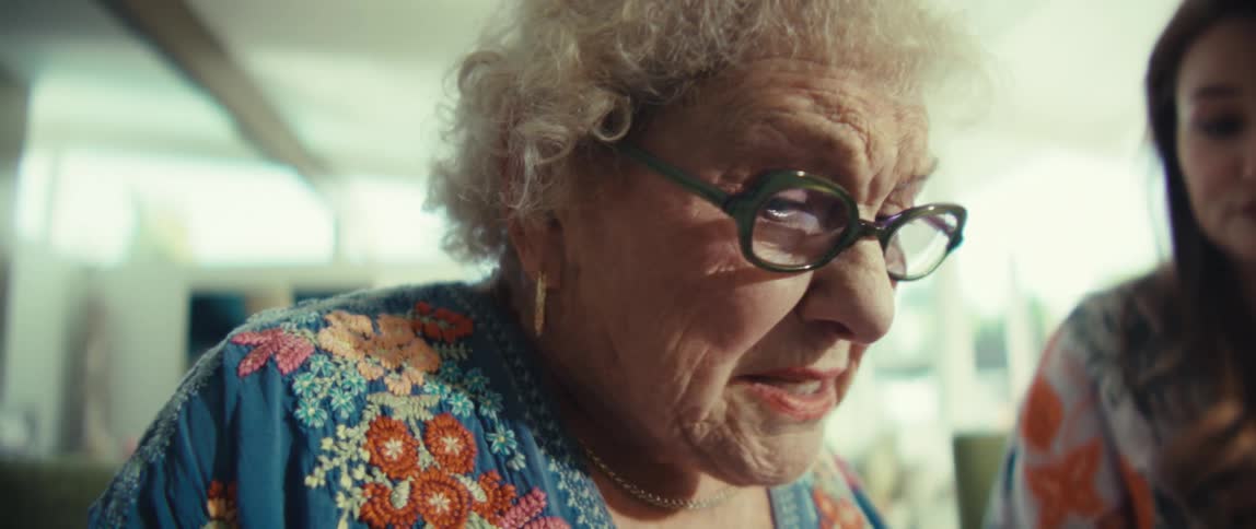 An elderly woman with curly white hair and glasses is sitting at a table wearing a blue embroidered shirt and working on a largebutton calculatornext to her a younger woman is partially visible holding a pen and appearing to assist with some pape