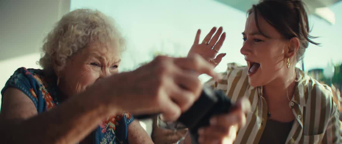 An older woman and a younger woman sit together indoors while excitedly playing a video game with controllers and cheering after winning