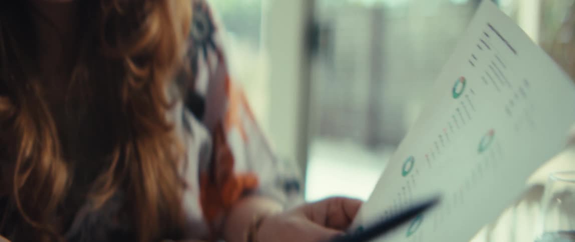 A close view shows a younger woman with long brown hair as she looks down and points at a document with financial charts using a pen