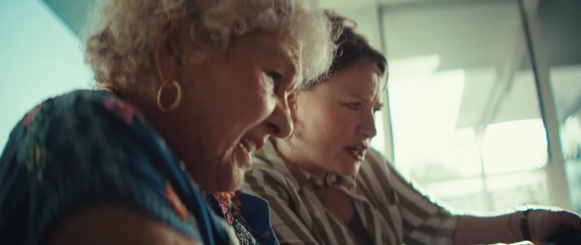 An elderly woman and a younger woman sit closely together on a couch focused intensely as they play a video game with handheld controllers the scene captures a playful bond between the two highlighted by bright sunlight and their expressive reactions t