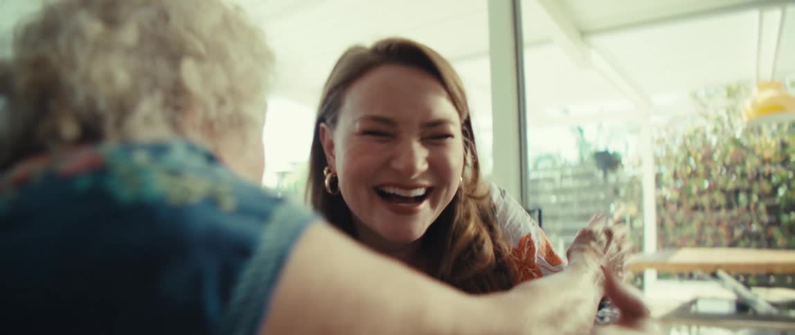 Two women looking at a screen with the young woman explaining something to the elderly woman both become excited hugging and smiling in the living room at midday