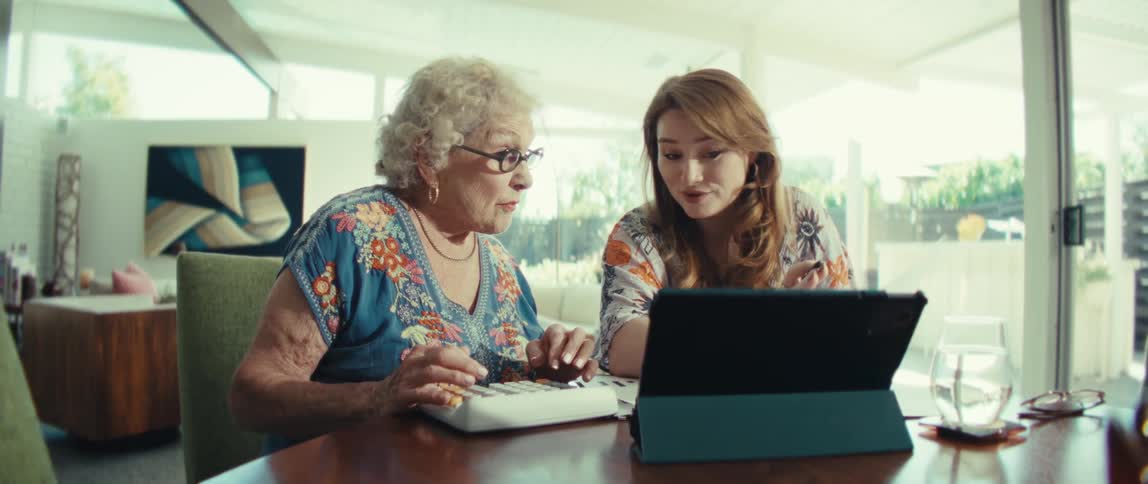 An elderly woman with glasses looks surprised while using a keyboard next to a young woman who holds a paper and points at a tablet screen on a wooden table in a bright modern room with large windows