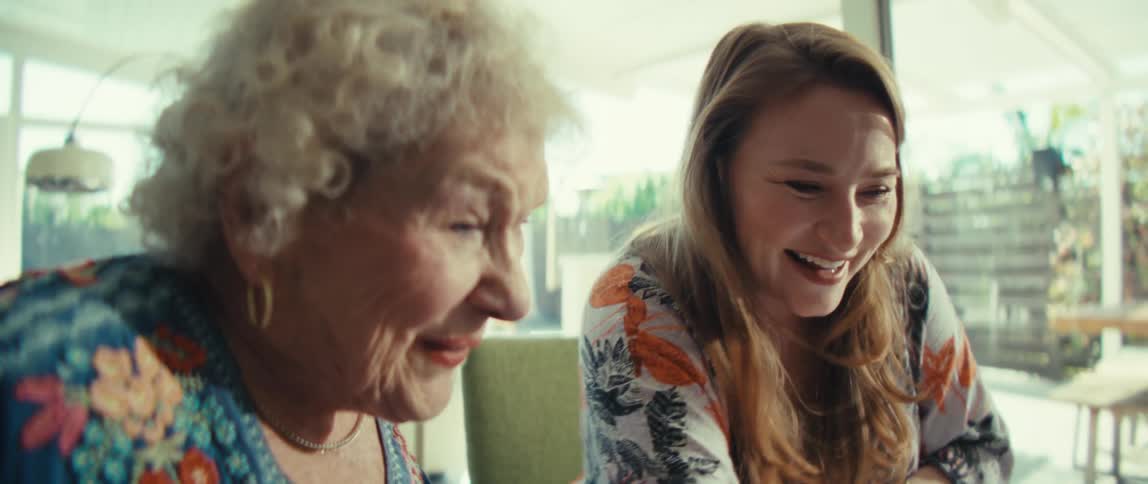 An older woman with curly gray hair and a younger woman wearing a floral shirt laugh while hugging and then look at a tablet and a calculator on a wooden table inside a bright house