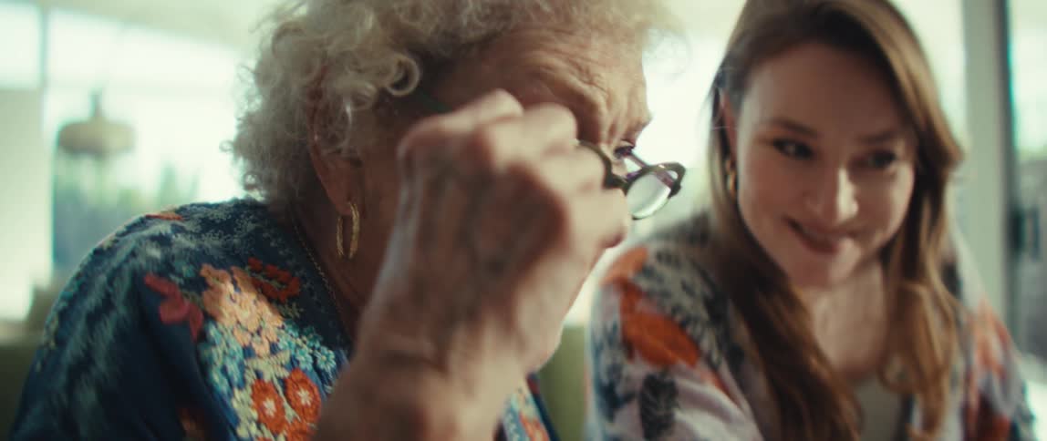 Two women sit together indoors where an elderly woman with white hair and glasses points at a tablet screen while a younger woman watches closely as they focus on the digital content in a brightly lit room