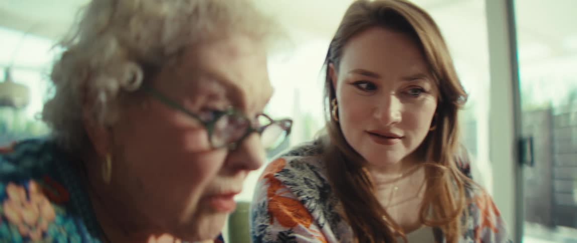 An elderly woman wearing glasses punching a calculator while looking at a tablet screen being guided by a young woman sitting beside her in the living room at midday