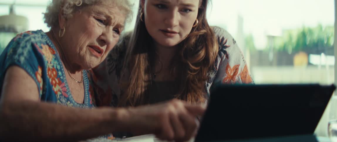 An elderly woman with white curly hair and a younger woman sit together at a table and look at a tablet with concerned expressions while the younger woman speaks and points at the screen in a bright room