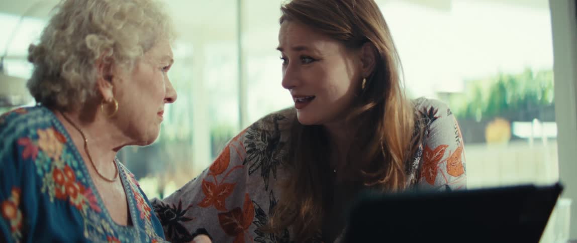 A younger woman wearing a floral shirt embraces an elderly woman while they sit together indoors with a tablet on the table and look at each other with sad expressions