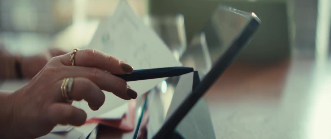 A woman wearing gold rings holds a pen and reviews a document containing various colorful pie charts and datashe smiles while looking at the information and pointing toward a nearby laptop screen