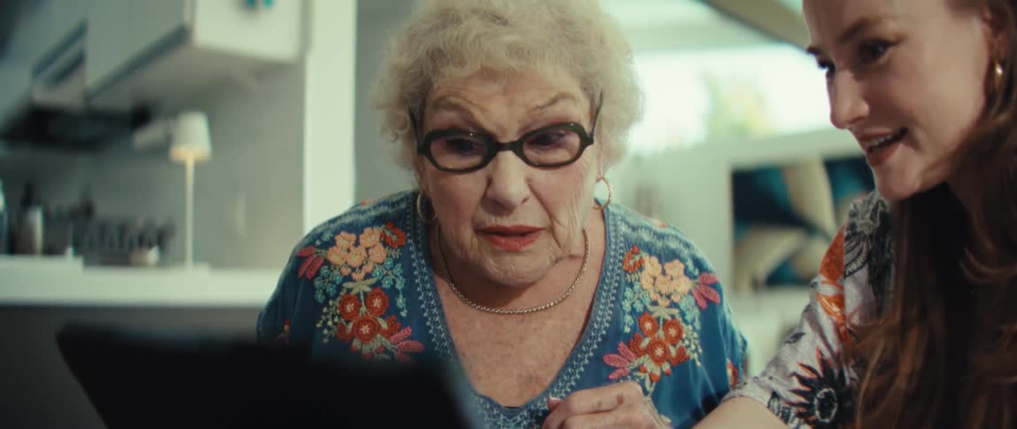 An elderly woman with white curly hair puts on black glasses and looks at a tablet screen with a surprised expression while a younger woman with long brown hair points at the device and talks to her in a bright room