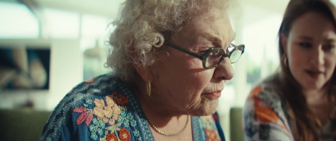 An elderly woman with curly white hair puts on her glasses while sitting next to a smiling younger woman and they are both interacting with a tablet computer in a bright living room