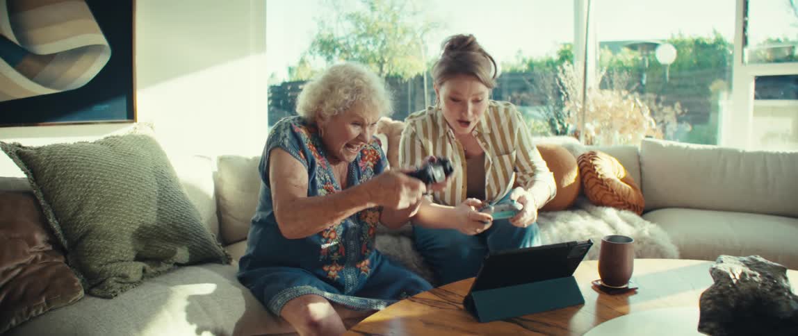 Woman sitting on a couch playing video games on a tablet computer with her granddaughter as they move the controllers in excitement