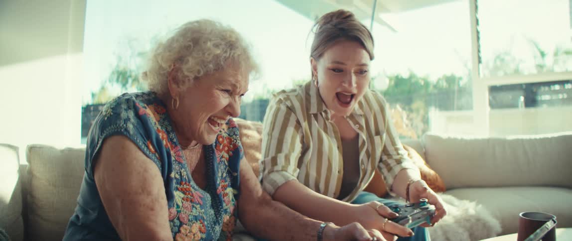 An elderly woman and a younger woman sit sidebyside on a sunlit couch gripping video game controllers with intense focus and excitementthey share a moment of highenergy bonding as they laugh and shout while competing or playing together on a table