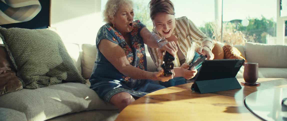 An elderly woman and a younger woman sit together indoors and use black controllers to play a video game with excited expressions while sunlight shines into the room from a nearby window