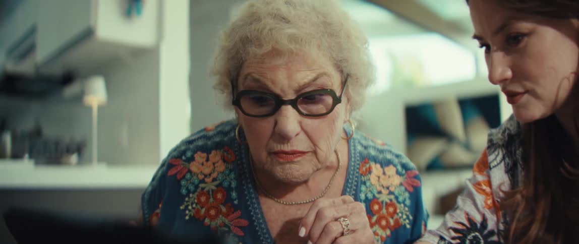 An elderly woman wearing a blue floral shirt puts on her blackrimmed glasses to get a better look at a laptop screena younger woman sits beside her pointing toward the screen as they both focus on the content together