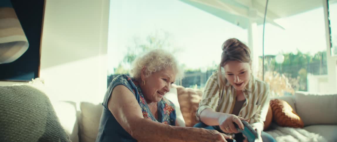 Two women one elderly and one young sitting on a couch in the living room at midday holding controllers and playing a video game having fun