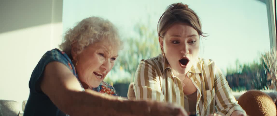 An elderly woman and a younger woman sit sidebyside leaning forward with intense focus as they play a video game togetherthe pair shifts between expressions of shock concentration and pure joy while gripped by the action on their controllers