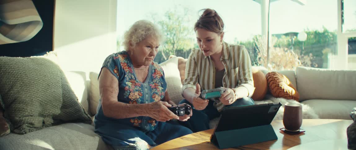 An elderly woman and a young woman sit on a sofa focused on playing a video game together using handheld controllers while a tablet sits on the wooden coffee table in a bright living room filled with soft pillows and natural sunlight