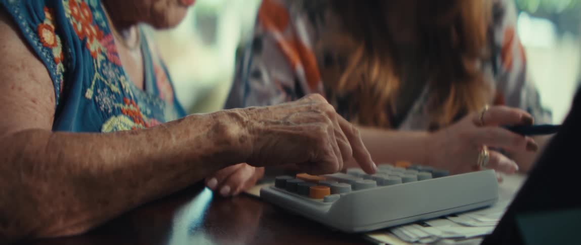 Two women sitting in front of a computer with the elderly woman using a calculator while the younger woman supervises