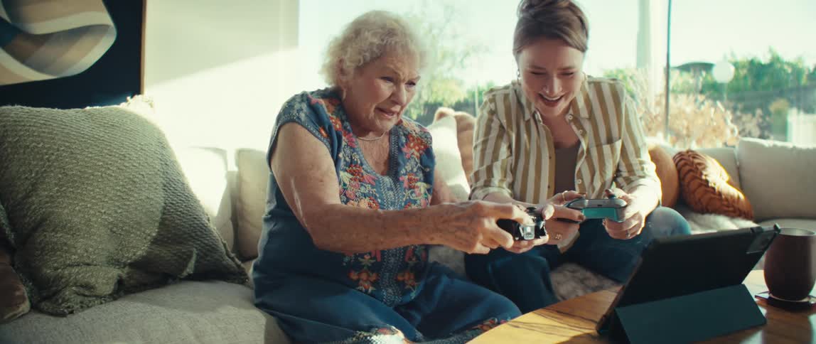 An elderly woman and a younger woman sit together indoors and use black controllers to play a video game with excited expressions while sunlight shines into the room from a nearby window
