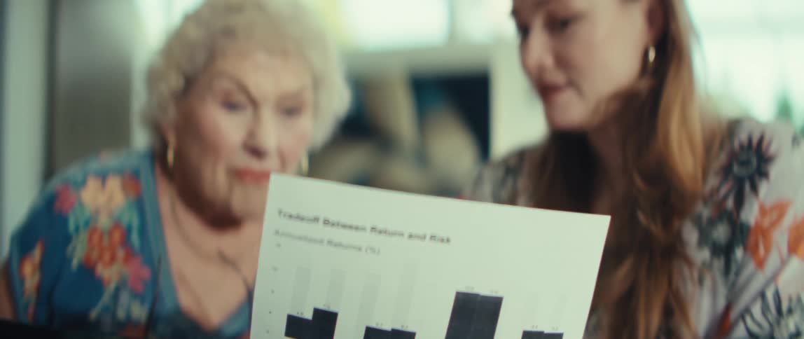 A younger woman explains a financial document featuring bar charts to an older woman the younger woman points to specific data while the older woman listens intently and adjusts her glasses