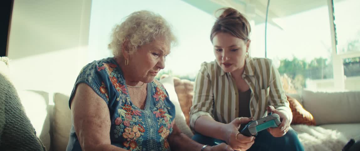 A younger woman and an elderly woman sit indoors on a sofa while the younger woman holds a blue game controller and explains how to use it to the older woman who holds a black controller