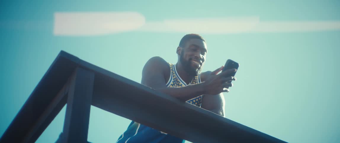A man with a black beard wearing a blue and yellow patterned basketball jersey leans over a wooden railing and smiles while using a smartphone against a clear blue sky on a bright day