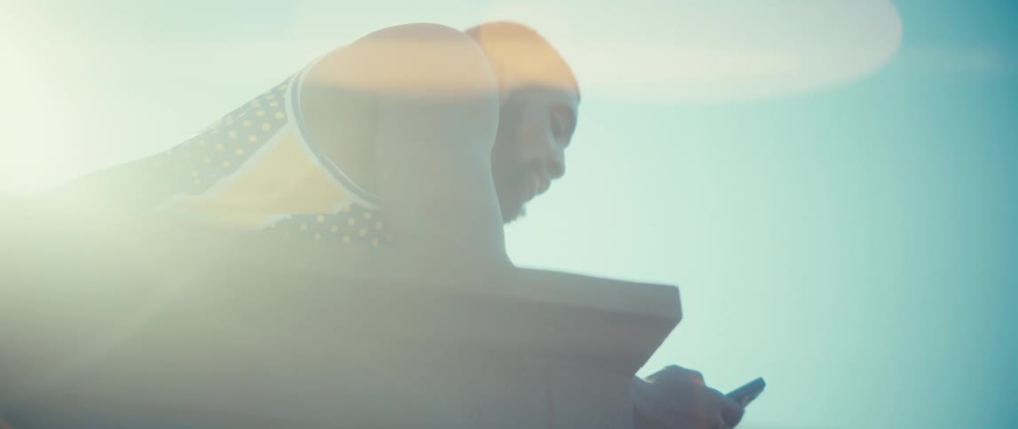 A man in athletic gear leans against a wooden railing under a bright hazy sky he looks down at a smartphone in his hands while sunlight creates a strong lens flare across the frame