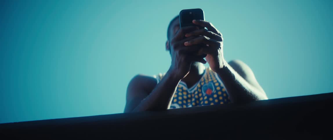 A man with a black beard wearing a blue and yellow patterned basketball jersey leans over a railing and uses a smartphone against a clear blue sky on a bright day