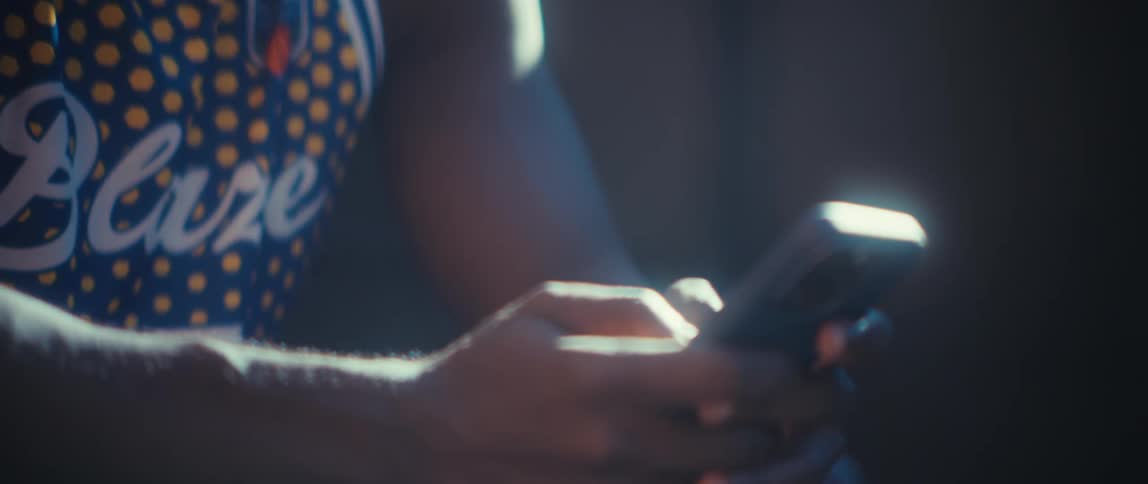 A young man wearing a jersey scrolling through his phone while seated outdoors under the sun