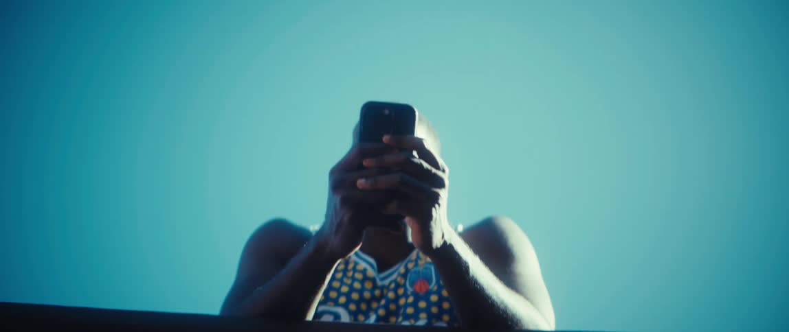 A black man in a patterned athletic top looks down at a smartphone from a low angle silhouetted against a vast cloudless blue sky