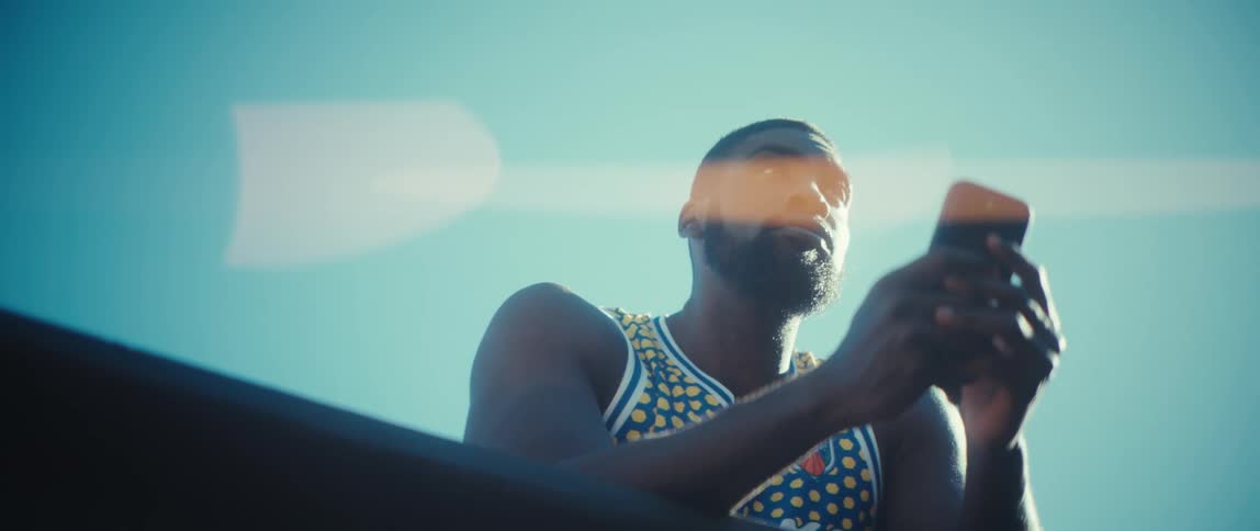 A black man wearing a yellow and blue basketball jersey leans against a metal railing while holding a smartphone under a bright clear sky with prominent lens flare