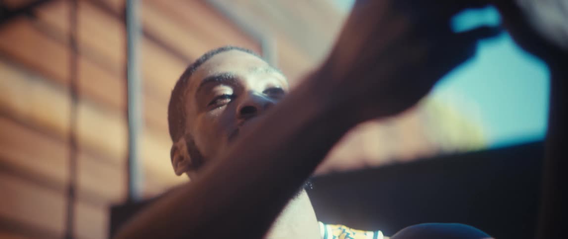 A man with a black beard wearing a basketball jersey looks upward while using a smartphone standing outdoors on a bright sunny day