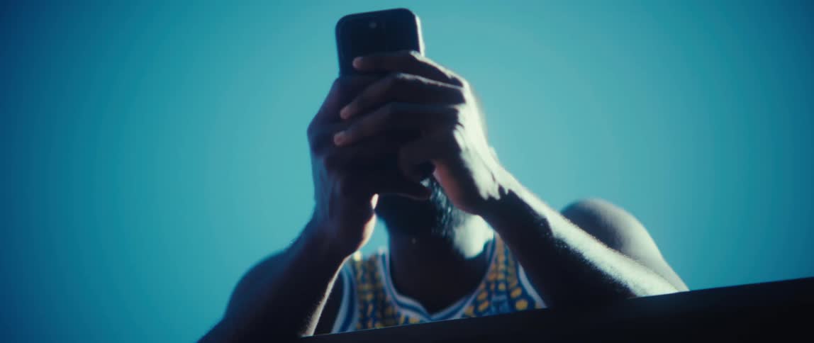 A man with a black beard wearing a blue and yellow patterned basketball jersey leans over a wooden railing and looks at his smartphone while standing outdoors against a clear blue sky on a bright day