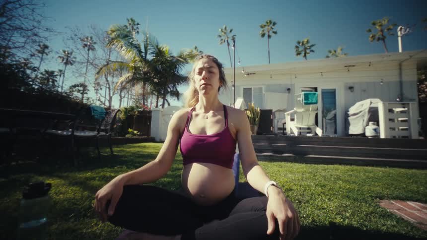 A pregnant woman sits cross legged on a yoga mat meditating in a sunny backyard