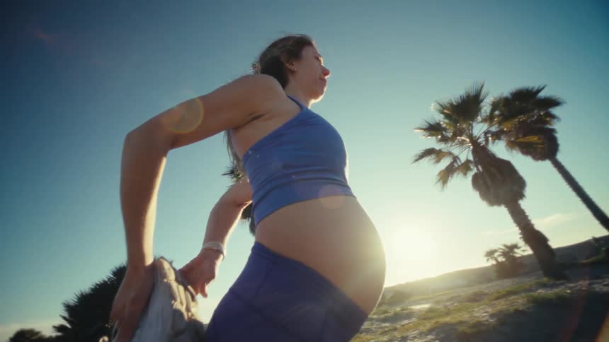 A pregnant woman stretching on a sandy beach with palm trees under a sunny sky