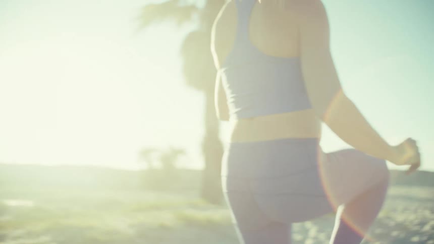 A view from the back shows a pregnant woman wearing a blue sports bra and blue leggings as she runs toward palm trees on a beach during sunset